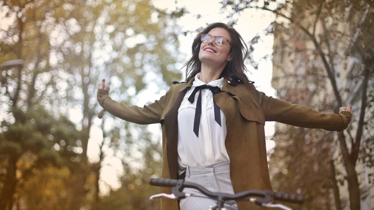 Low angle of cheerful female wearing old fashioned jacket and blouse sitting on bike with closed eyes and outstretched arms while enjoying weekend